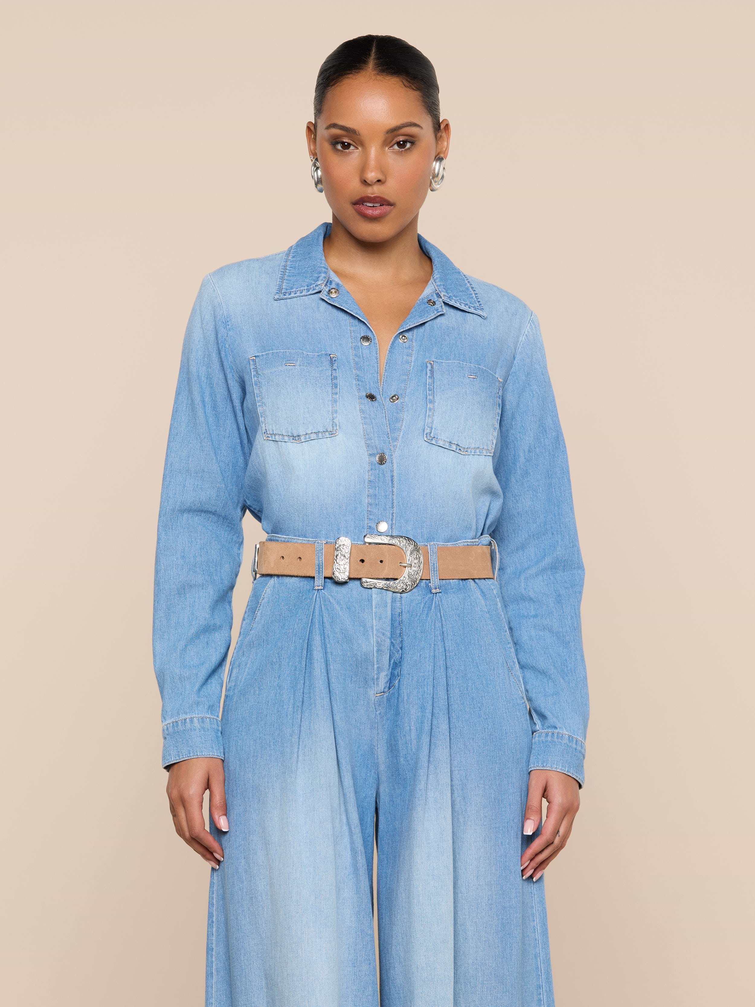 A woman stands facing forward in the Isla Shirt, styled with a beige belt and silver earrings, against a plain beige background.