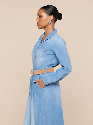 A woman in profile stands against a beige background, wearing the Isla Shirt in light blue denim with a beige belt, large silver hoop earrings, her hair in a neat bun, and her hands tucked into her pockets.