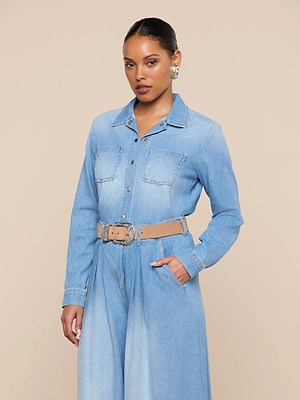 A woman with hoop earrings and slicked-back hair models the Isla Shirt, a lightweight light blue denim button-up with chest pockets and a beige belt, standing against a plain beige background, one hand in her pocket.