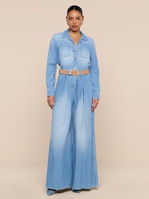 A woman poses against a beige backdrop wearing the Isla Shirt—a lightweight, light blue denim top with button front and flap chest pockets—paired with wide-leg pants, a tan belt, and small hoop earrings.