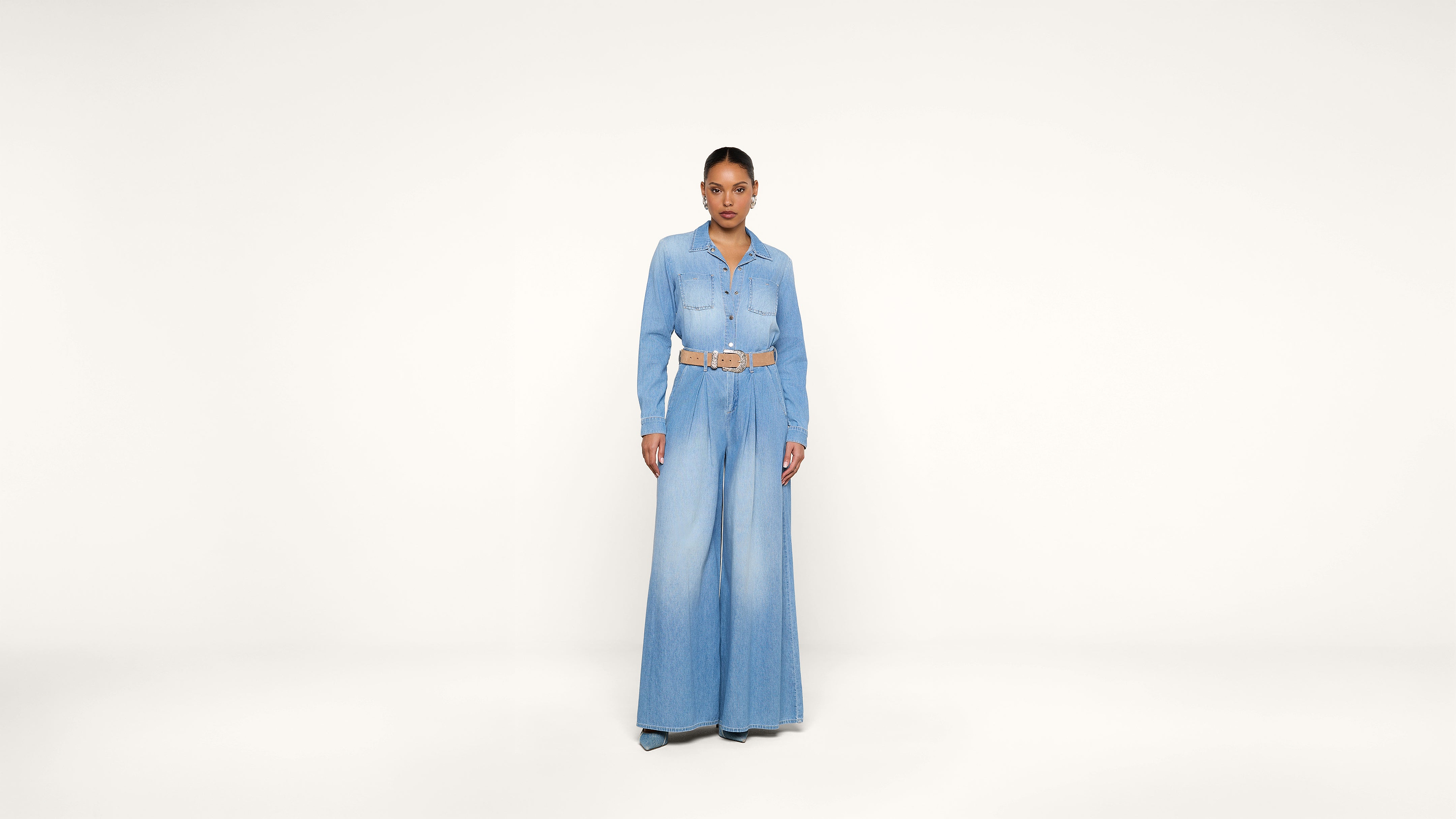 A woman stands facing forward wearing a light blue denim jumpsuit with wide-leg pants and a tan belt, against a plain white background.