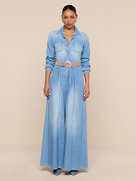 A woman poses against a beige background wearing the Isla Shirt, a lightweight, light blue denim top with long sleeves, styled with wide-leg pants, a beige belt, and teal high-heeled shoes.