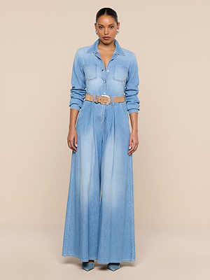 A woman poses against a beige background wearing the Isla Shirt, a lightweight, light blue denim top with long sleeves, styled with wide-leg pants, a beige belt, and teal high-heeled shoes.