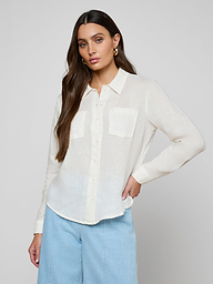 A woman wearing the Isla Linen Shirt—a white linen button-down with two front pockets—paired with light blue pants, stands against a plain light background with her hand on her hip.