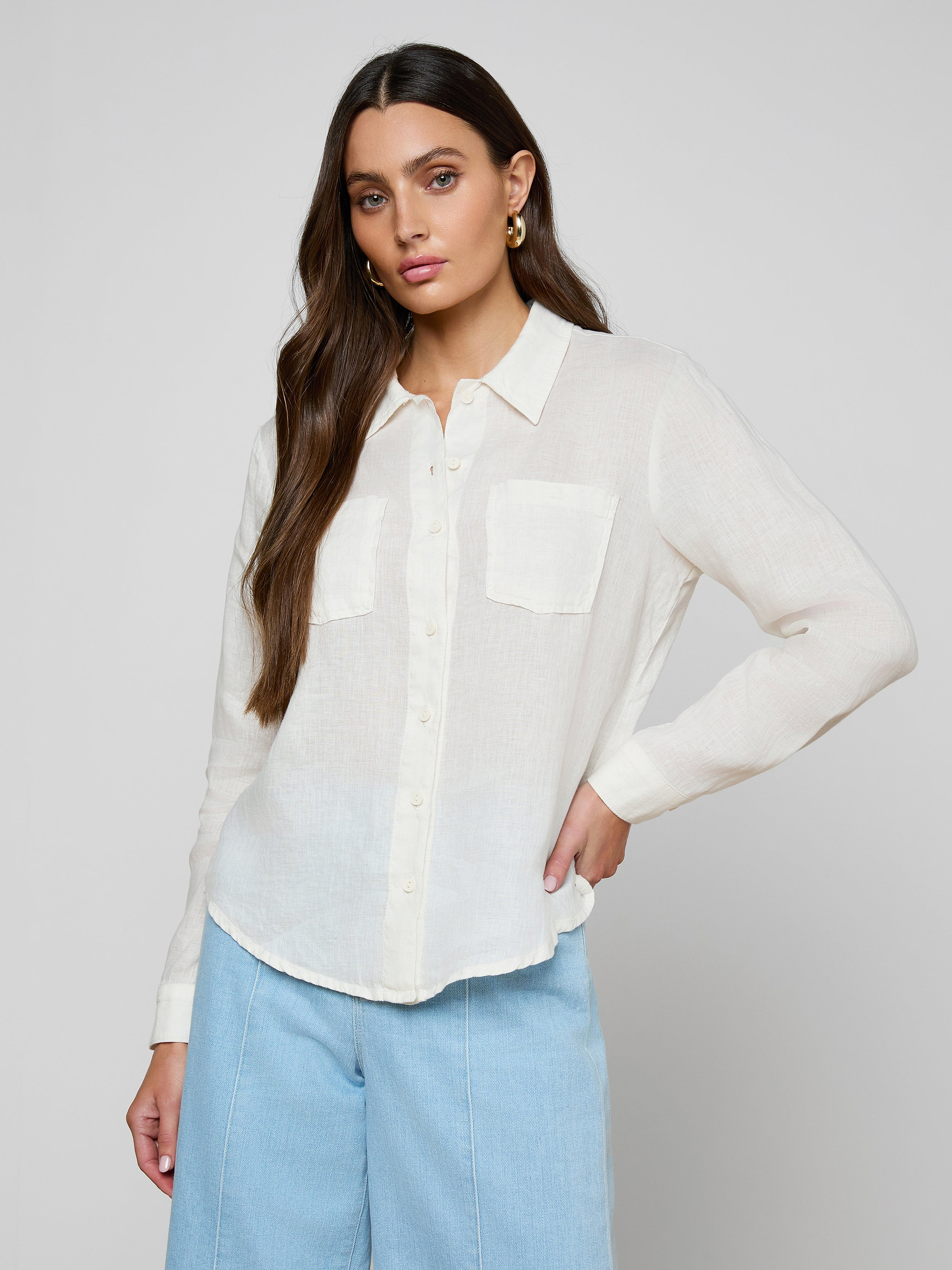 A woman wearing the Isla Linen Shirt—a white linen button-down with two front pockets—paired with light blue pants, stands against a plain light background with her hand on her hip.