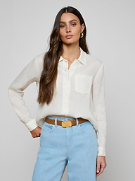 A woman with long brown hair wears the Isla Linen Shirt, a white button-down, paired with light blue high-waisted jeans as she stands with one hand on her hip against a plain background.
