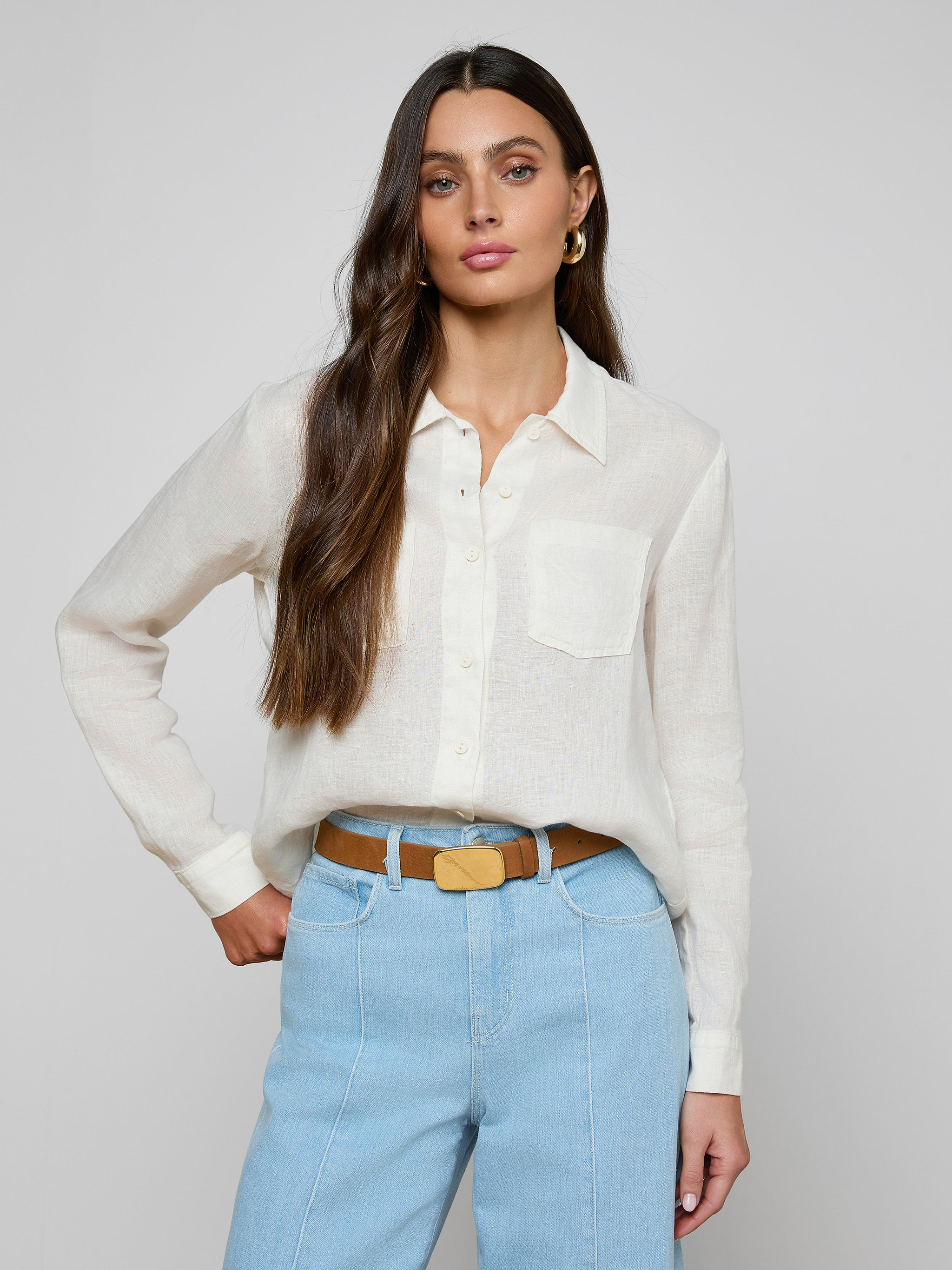 A woman with long brown hair wears the Isla Linen Shirt, a white button-down, paired with light blue high-waisted jeans as she stands with one hand on her hip against a plain background.