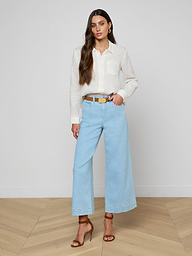 A woman stands against a plain white background, wearing the Isla Linen Shirt with long sleeves, paired with light blue wide-leg jeans, a brown belt, and brown high-heeled sandals. She has long brown hair and a neutral expression.