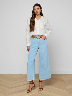 A woman with long brown hair and a neutral expression stands indoors against a plain wall, wearing the Isla Linen Shirt tucked into high-waisted, wide-leg light blue jeans, styled with a brown belt and heeled sandals.