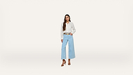 A woman stands against a plain white background, wearing a white blouse, light blue wide-leg jeans, brown belt, and brown heeled sandals, with long brown hair styled in loose waves.