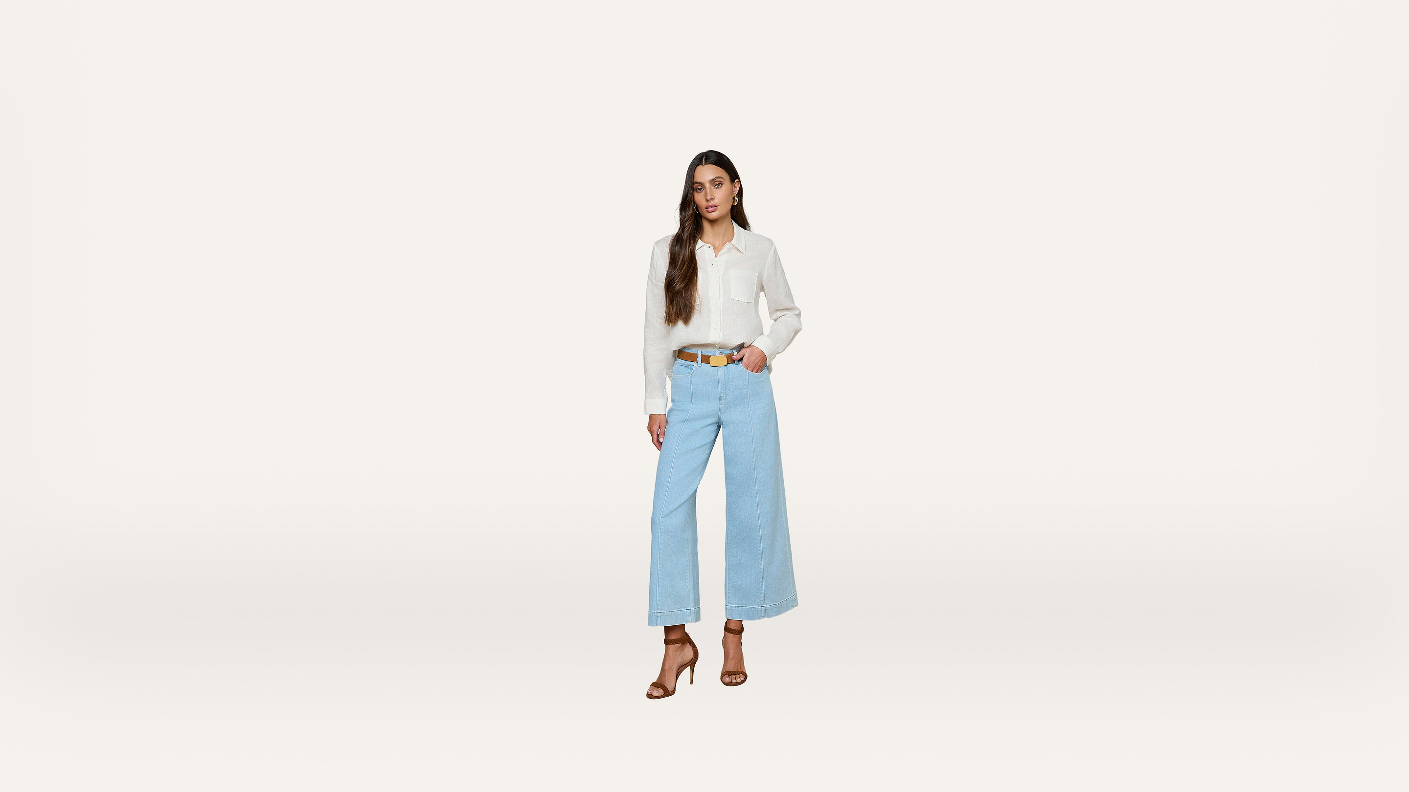 A woman stands against a plain white background, wearing a white blouse, light blue wide-leg jeans, brown belt, and brown heeled sandals, with long brown hair styled in loose waves.
