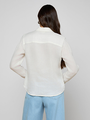 A person with long brown hair models the Isla Linen Shirt—a white button-down—paired with light blue jeans, standing with their back to the camera against a plain light gray backdrop.