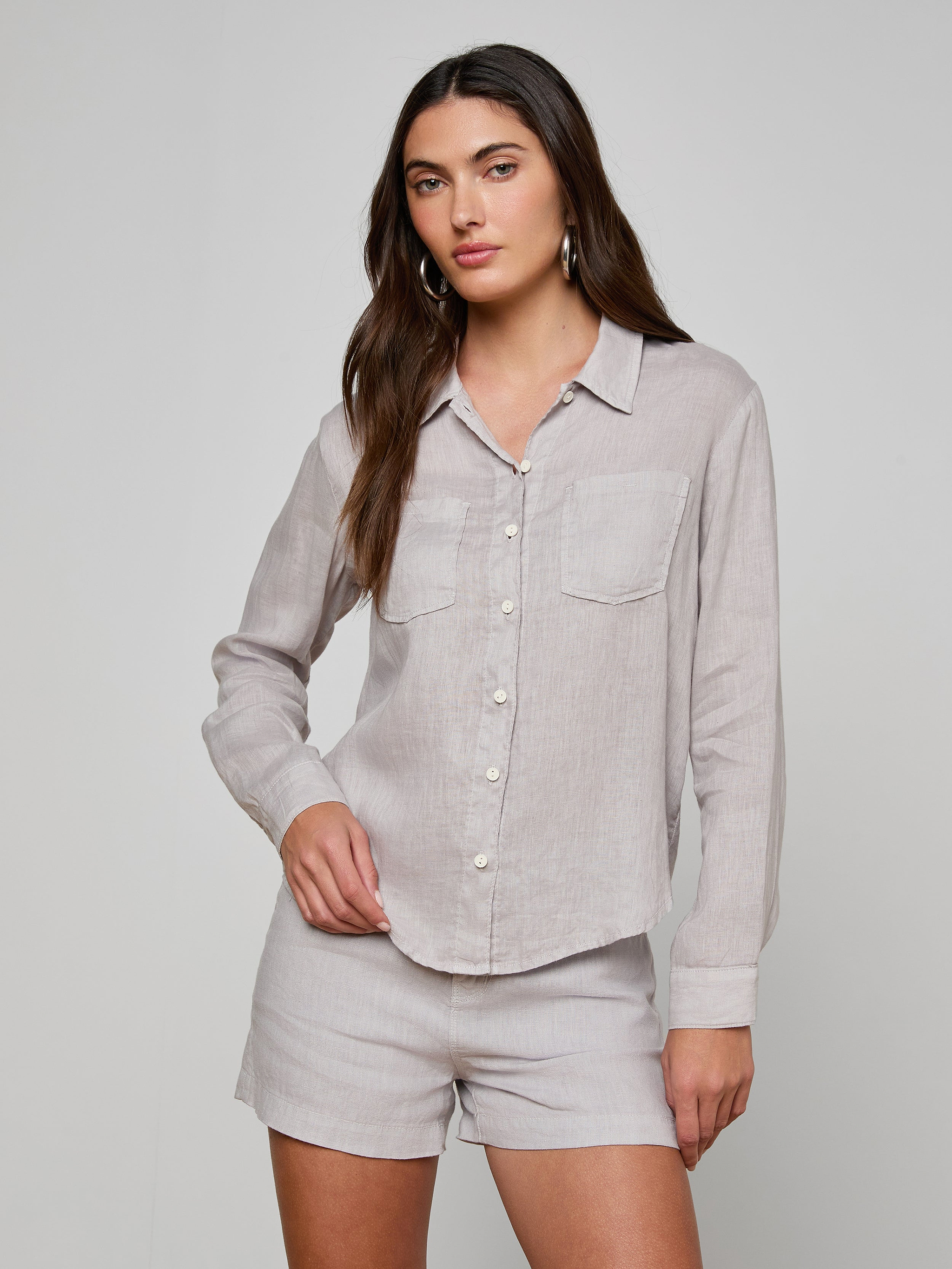 A woman stands against a plain background wearing the Isla Linen Shirt, a light grey, long-sleeve button-down with two front pockets, paired with matching shorts.