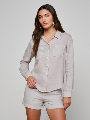 A woman with long brown hair wears the Isla Linen Shirt in light grey, featuring chest pockets, paired with matching shorts. She poses with one hand in her pocket against a plain, light background.