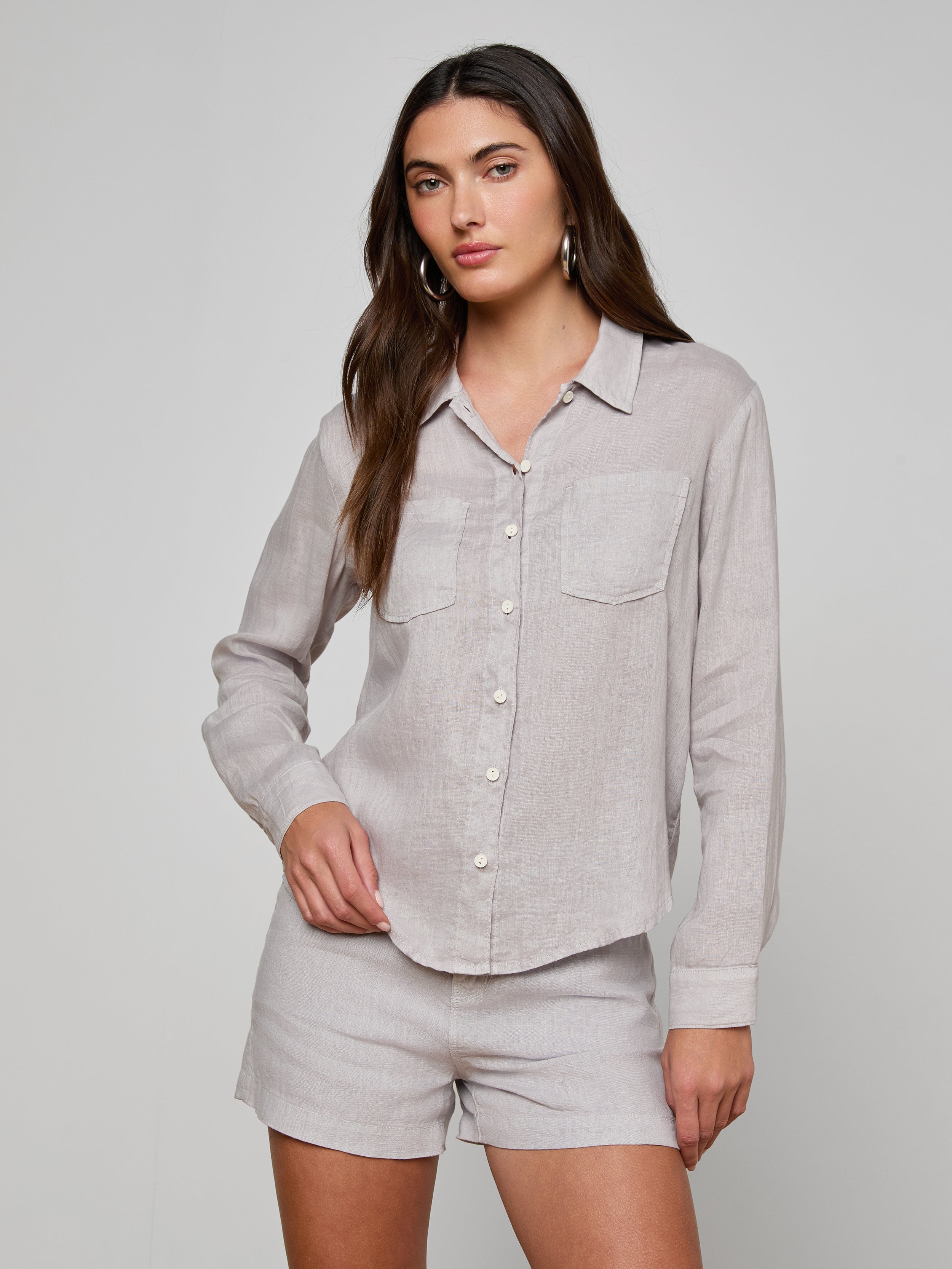 A woman stands against a plain background wearing the Isla Linen Shirt, a light grey, long-sleeve button-down with two front pockets, paired with matching shorts.
