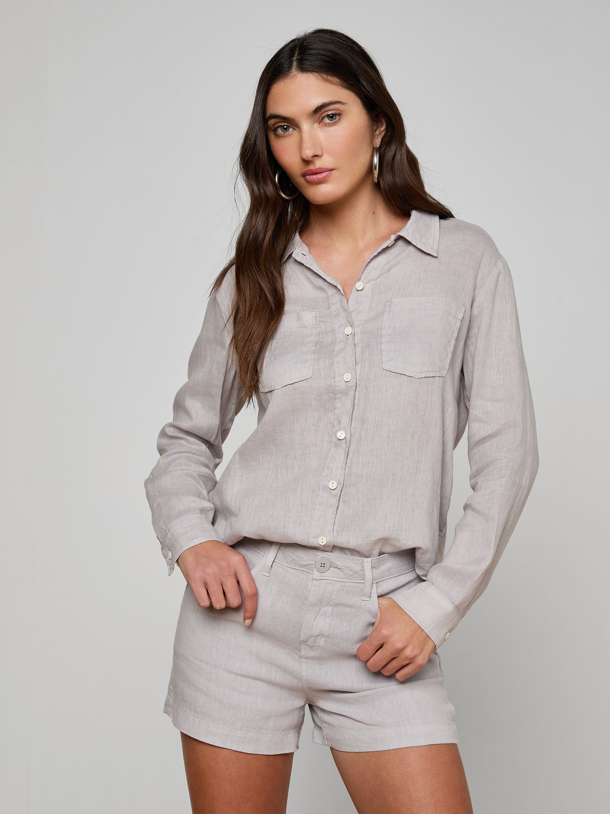 Wearing the Isla Linen Shirt in light grey, a woman with one hand in her pocket stands against a plain background; the button-down shirt features long sleeves and is paired with matching shorts.
