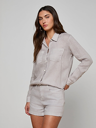 A woman with long brown hair wears the Isla Linen Shirt, a light beige button-down style with front pockets, paired with matching shorts. She stands against a plain, light background, looking toward the camera in a relaxed pose.