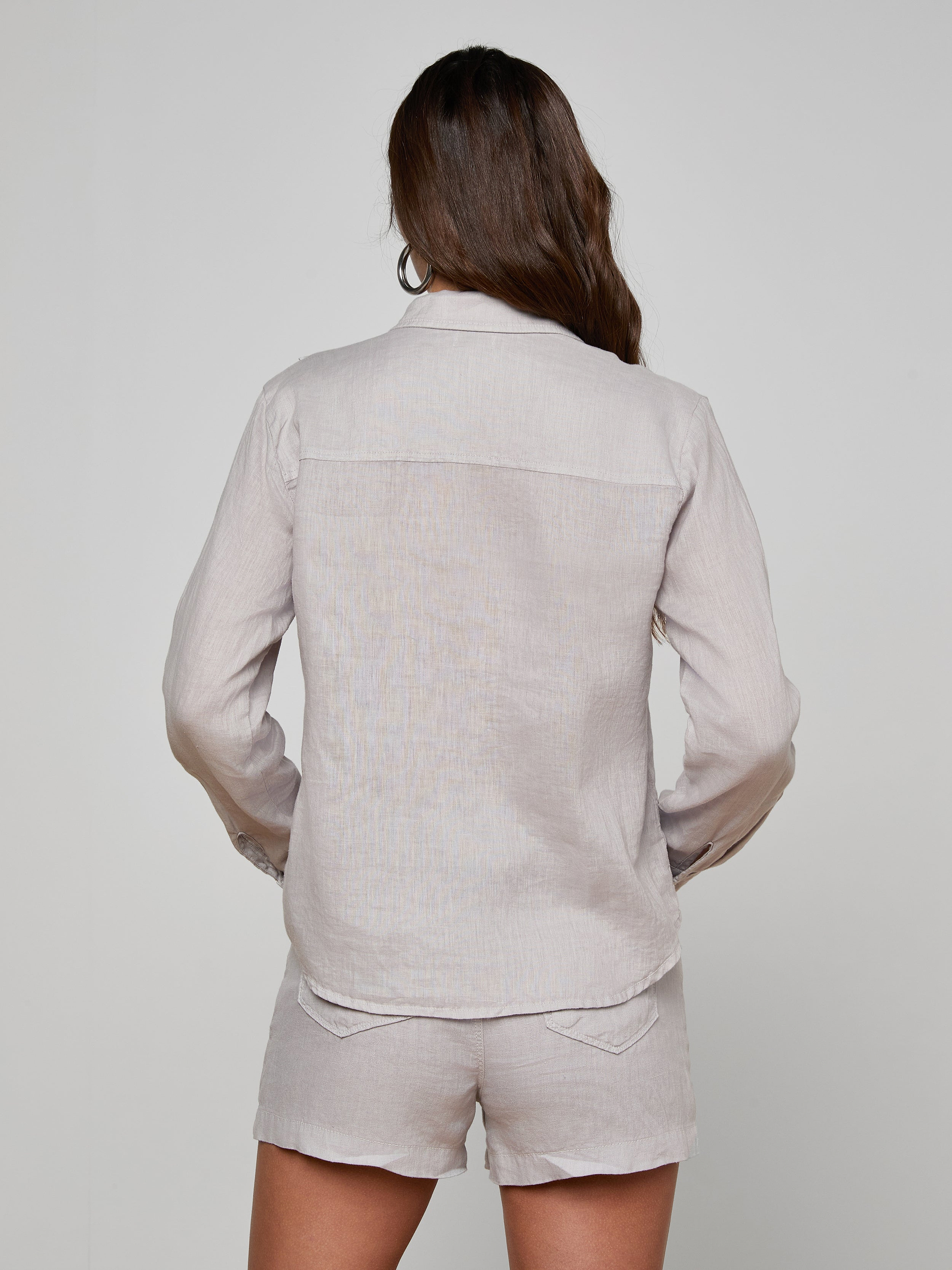 Wearing the Isla Linen Shirt, a woman with long brown hair is seen from behind in a light-colored ensemble, standing against a plain light background.