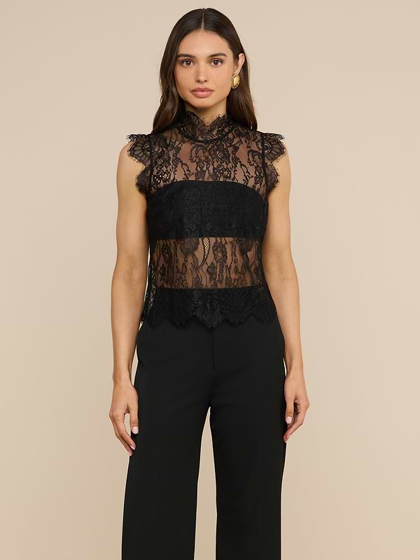 A woman stands against a plain light background, facing forward with her arms at her sides, wearing the Tours Lace Top— a sleeveless, sheer Chantilly lace design with a high neck—paired with black pants.