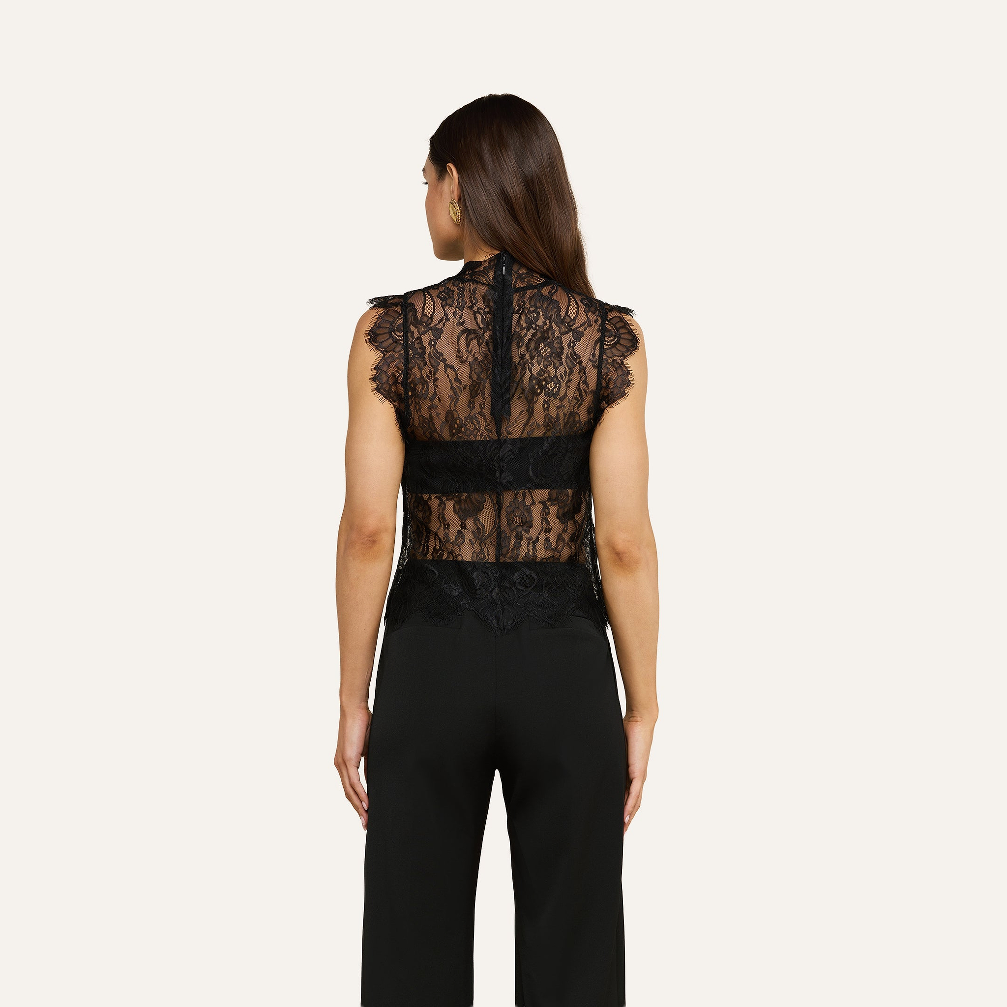 A woman with long brown hair is shown from the back against a plain light background, wearing the sleeveless, sheer Tours Lace Top paired with black pants.