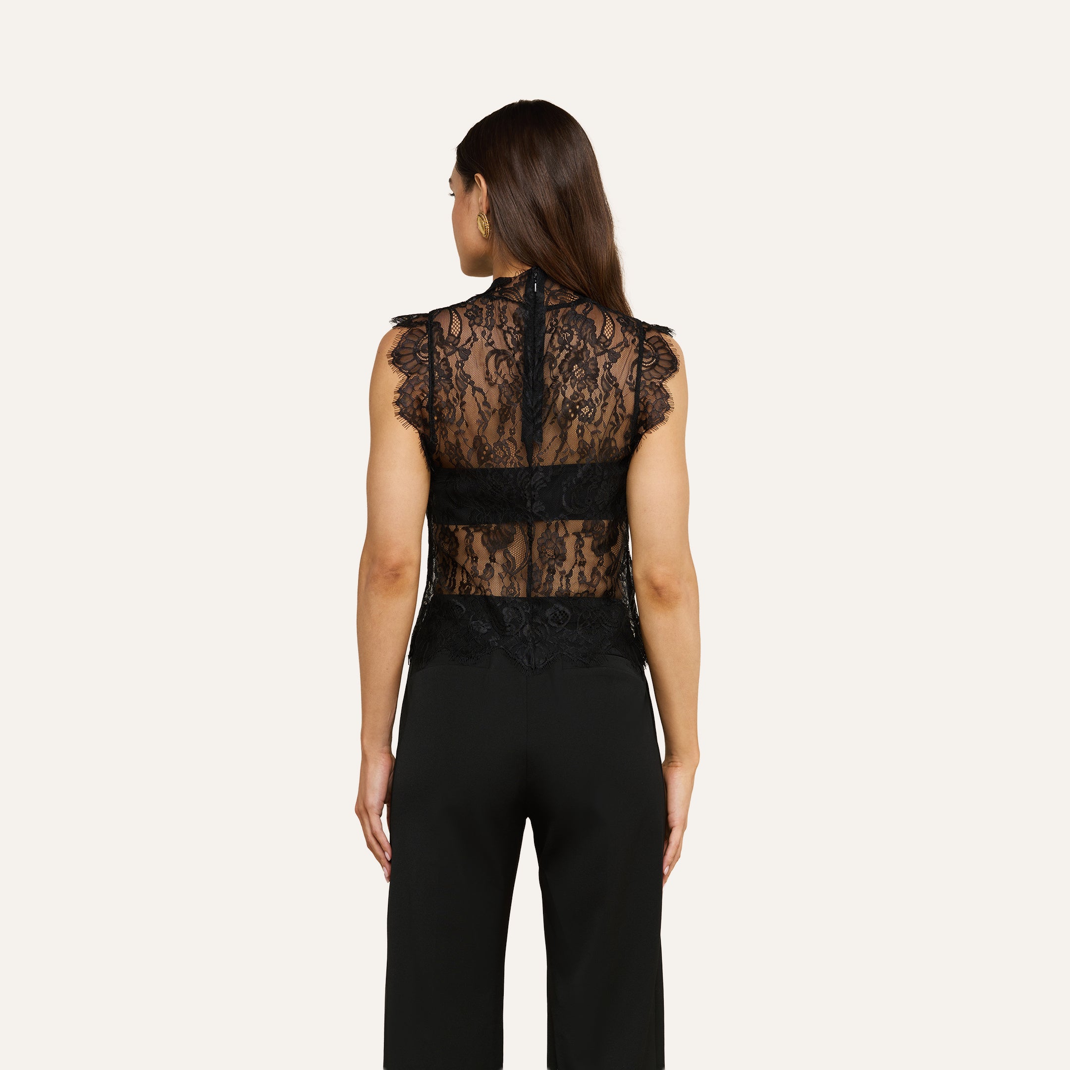 A woman with long brown hair is shown from the back against a plain light background, wearing the sleeveless, sheer Tours Lace Top paired with black pants.