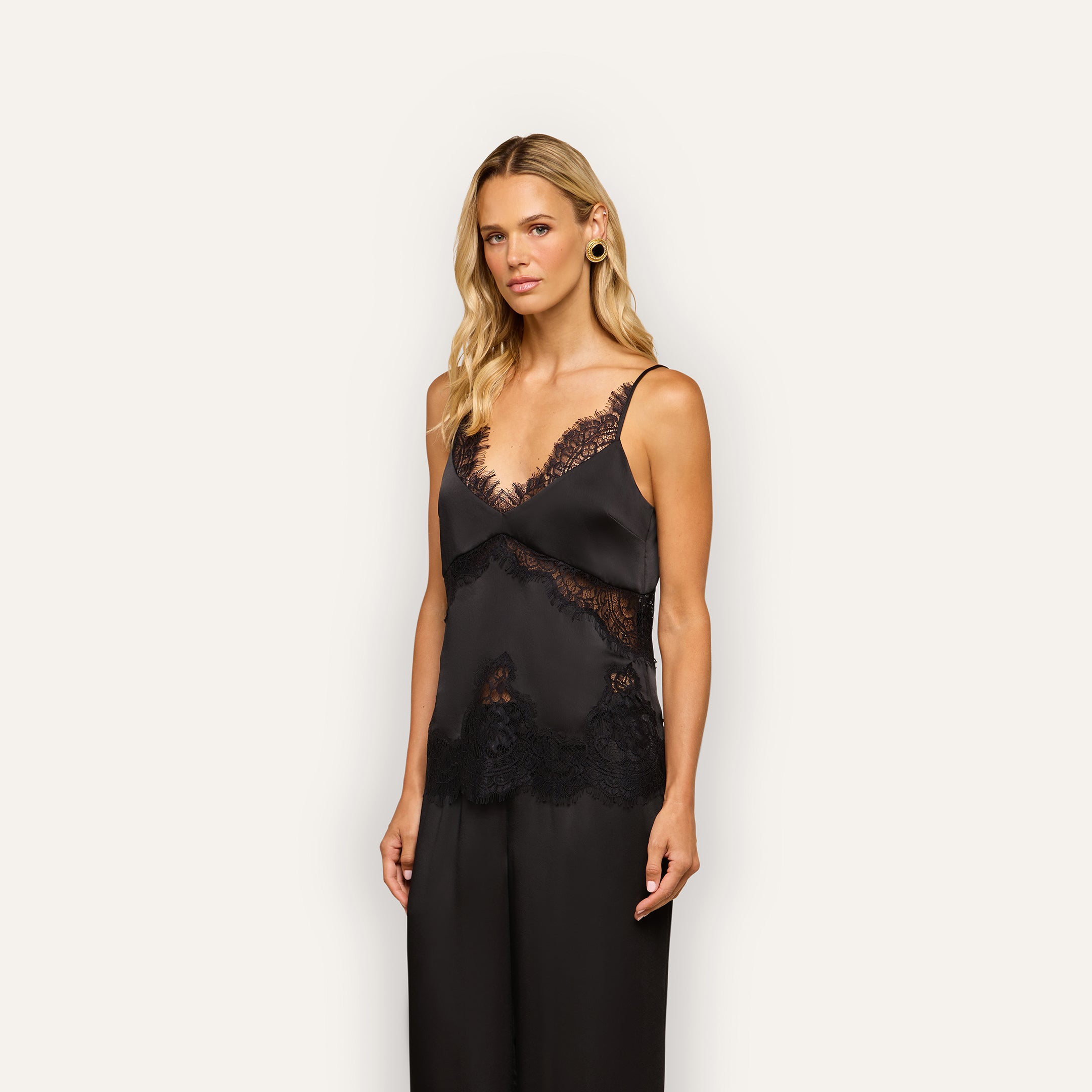 A woman with blonde hair wears the Mitsu Satin Lace Camisole and matching black pants, standing against a plain light background and looking toward the camera with a neutral expression.