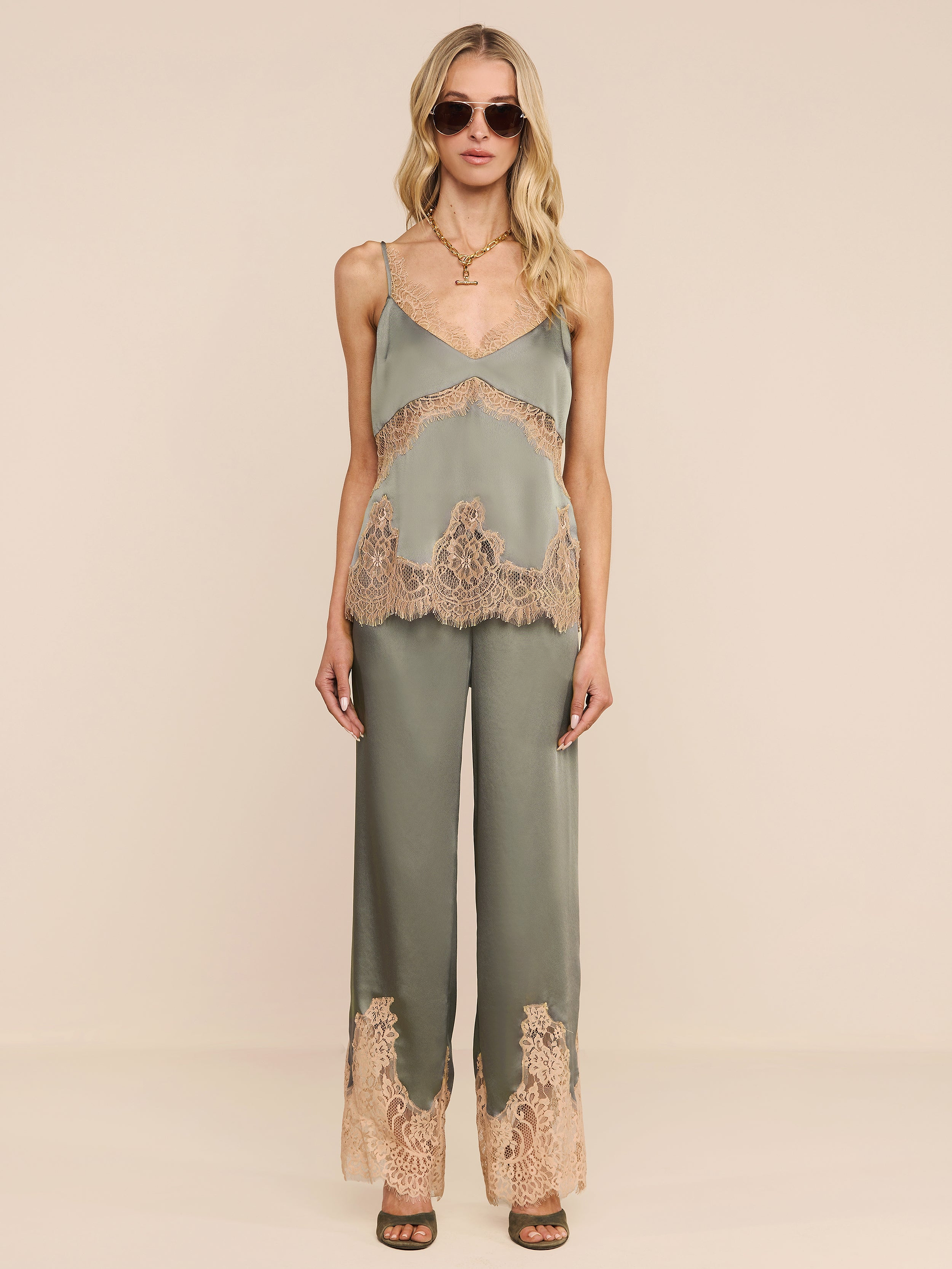 Una donna con occhiali da sole e la canotta in pizzo di seta Mitsu, un top verde salvia con spalline regolabili, abbinato a pantaloni a gamba larga con inserti in pizzo beige, si trova su uno sfondo beige. Ha lunghi capelli biondi ondulati e sandali con tacco marrone.