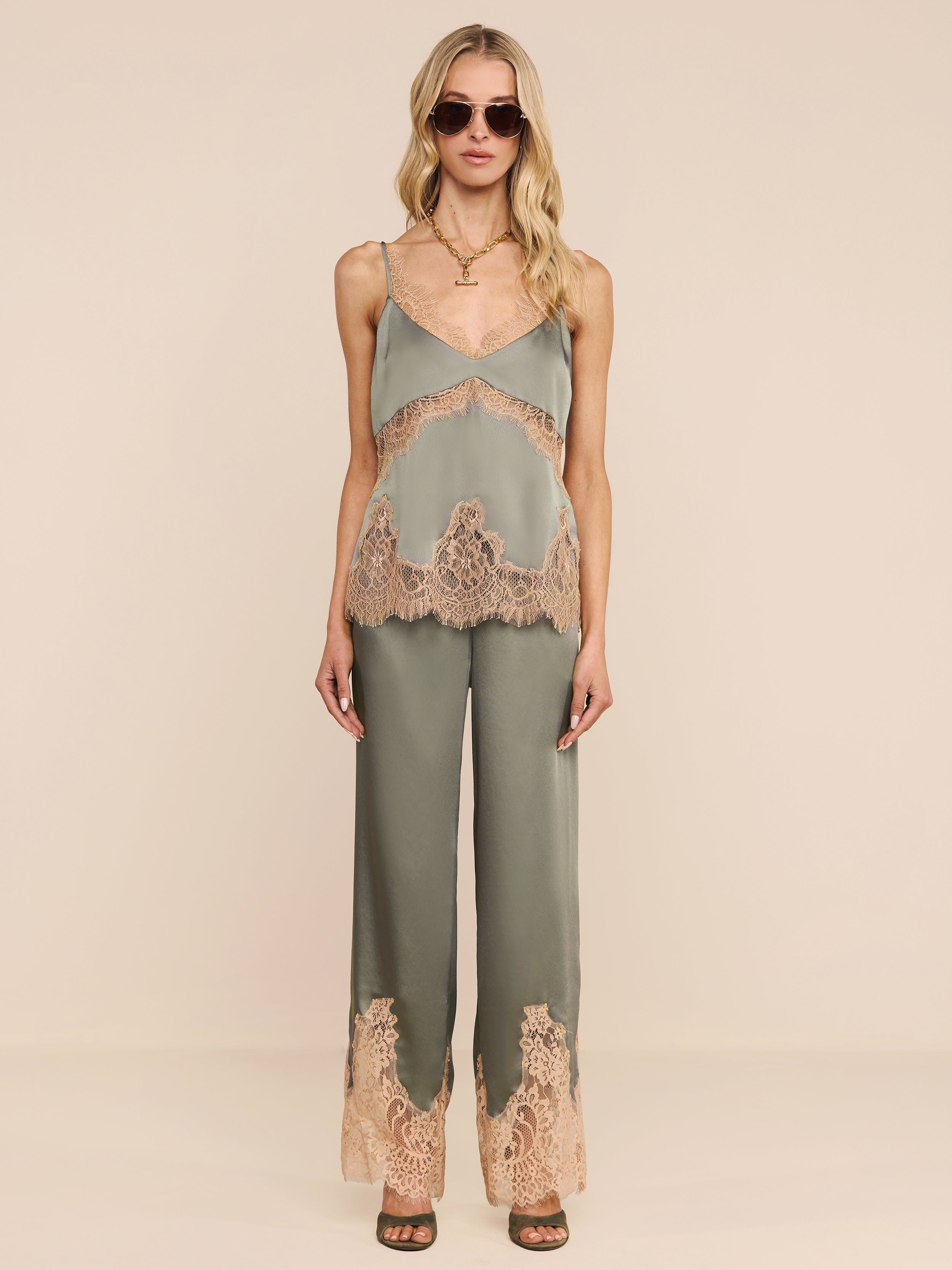 Una donna con occhiali da sole e la canotta in pizzo di seta Mitsu, un top verde salvia con spalline regolabili, abbinato a pantaloni a gamba larga con inserti in pizzo beige, si trova su uno sfondo beige. Ha lunghi capelli biondi ondulati e sandali con tacco marrone.