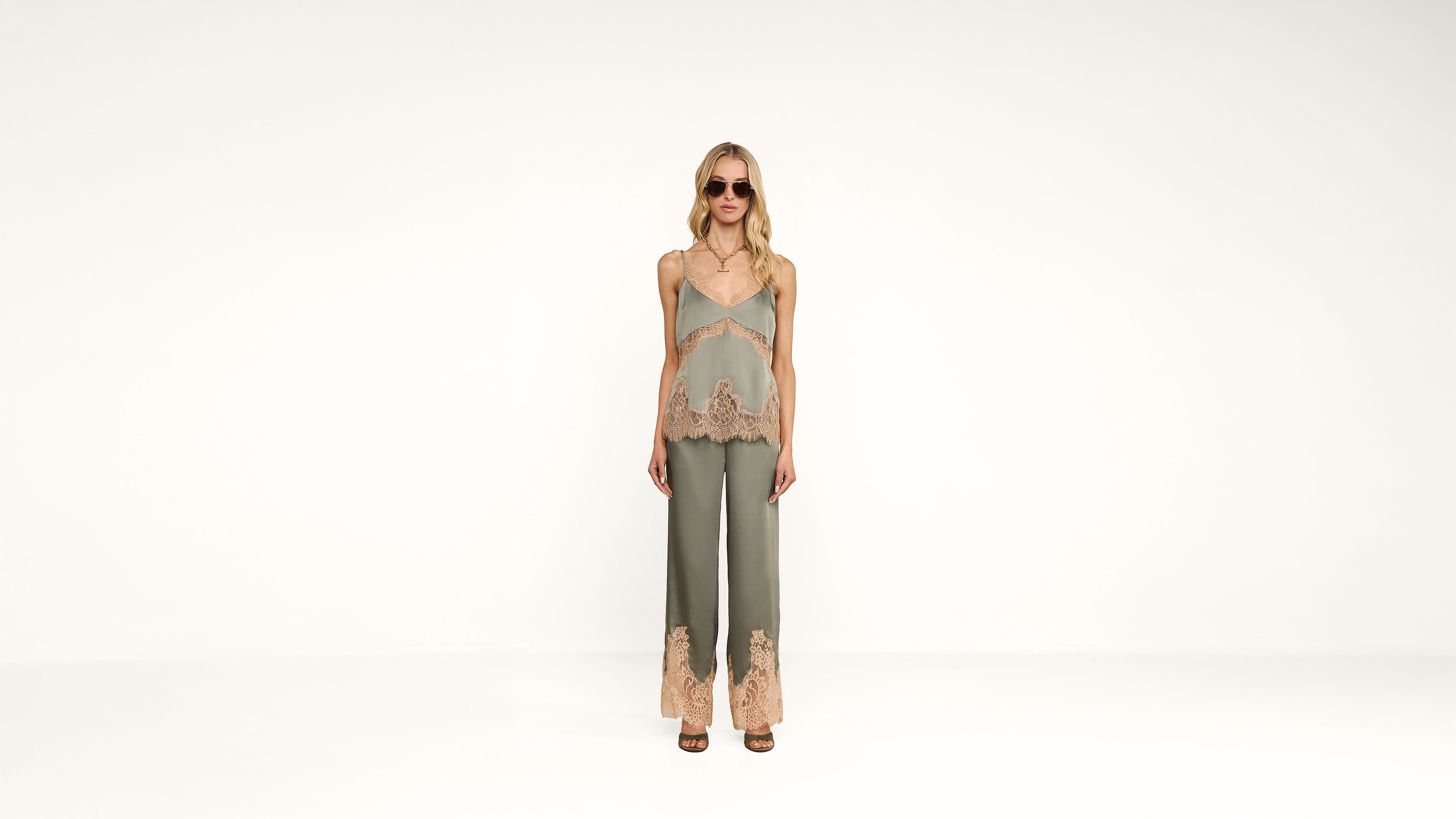 A woman wearing sunglasses, a green camisole top, and matching green pants with beige lace trim stands against a plain white background.