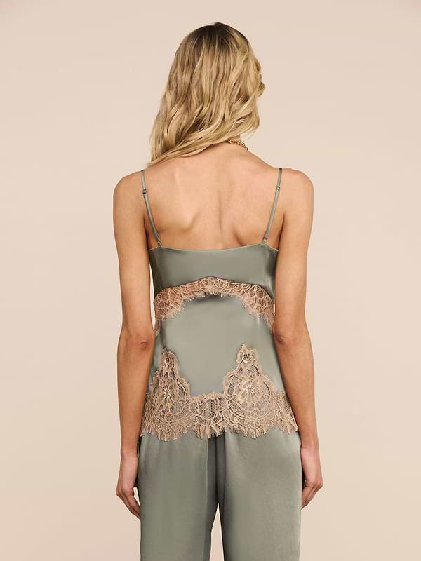 Una donna con capelli biondi ondulati, vista da dietro, indossa la canotta in pizzo di seta Mitsu in verde salvia con delicati inserti in pizzo e spalline regolabili, abbinata a pantaloni coordinati. Lo sfondo è un semplice beige chiaro.