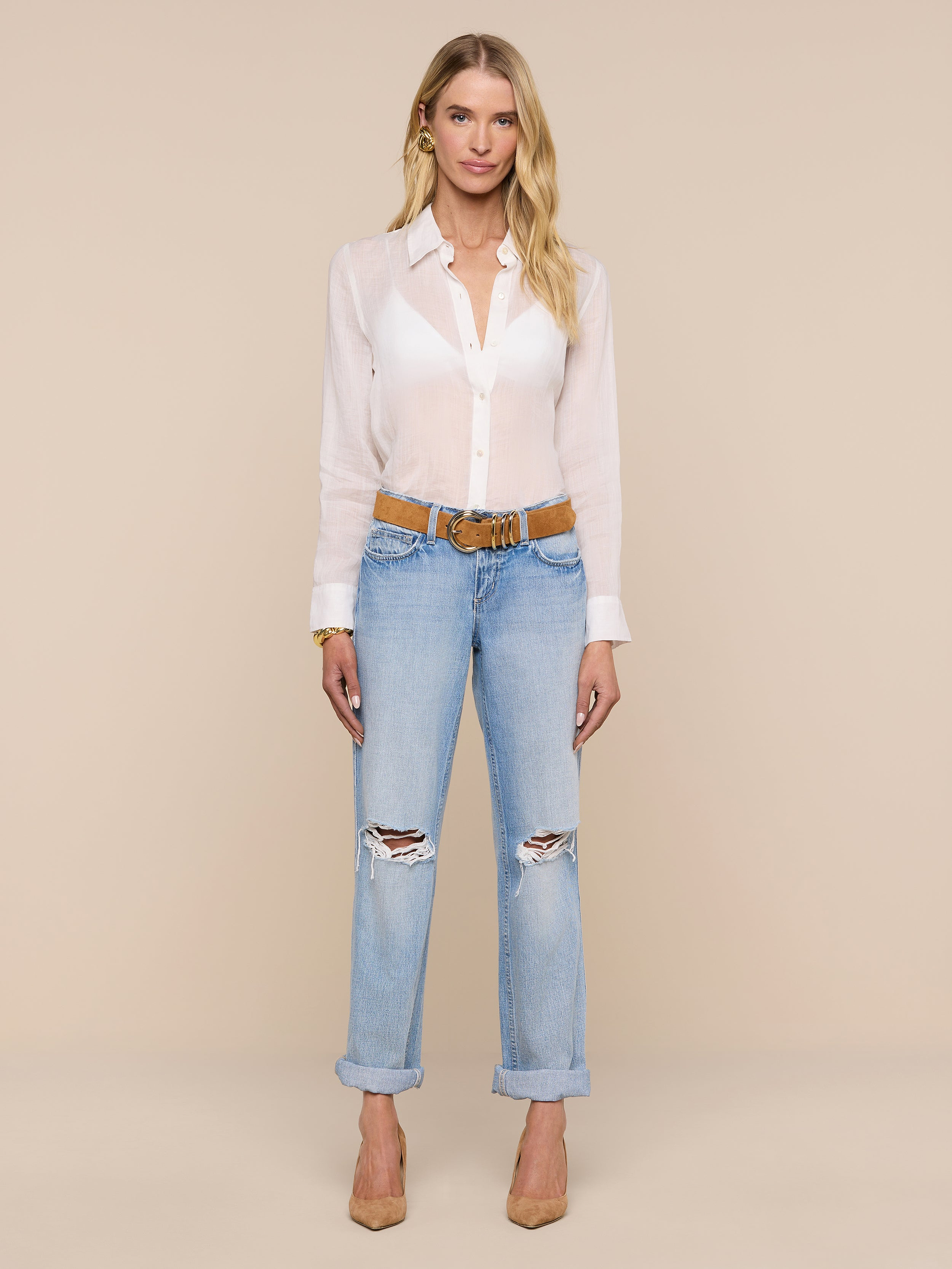 A woman stands facing forward, wearing the Ellery Blouse—a white sheer ramie shirt—paired with ripped light blue jeans with cuffs, a brown belt, and tan heels against a plain light background.