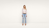 A woman stands in front of a plain white background, wearing a sheer white blouse, light blue ripped jeans, a brown belt, and tan heels. She has long blonde hair and is facing forward with a neutral expression.