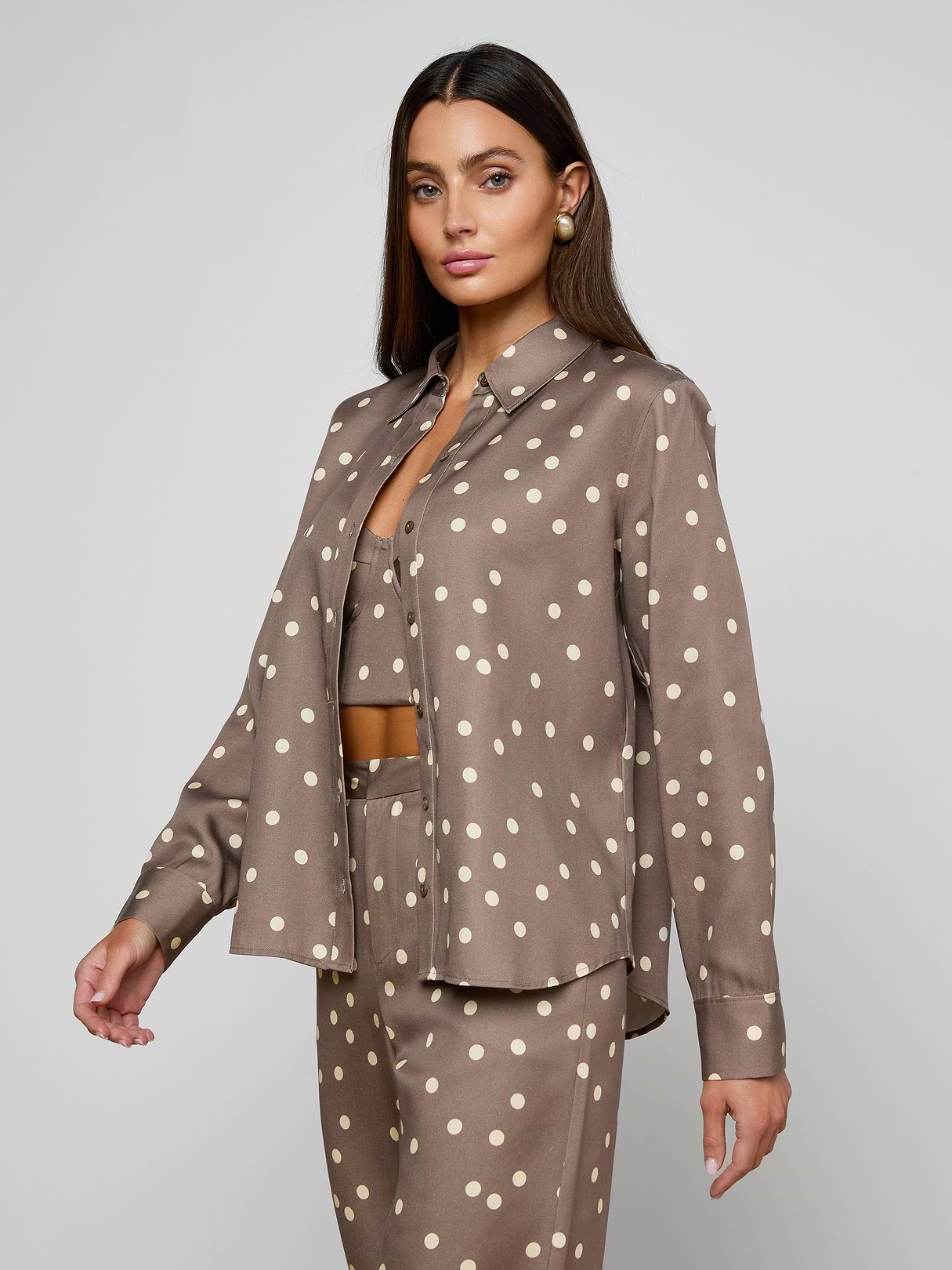 Woman wearing the Ellery Button-Down Shirt in cardamom brown with white polka dots, paired with matching long-sleeve pants and a coordinating crop top, standing against a plain light background.