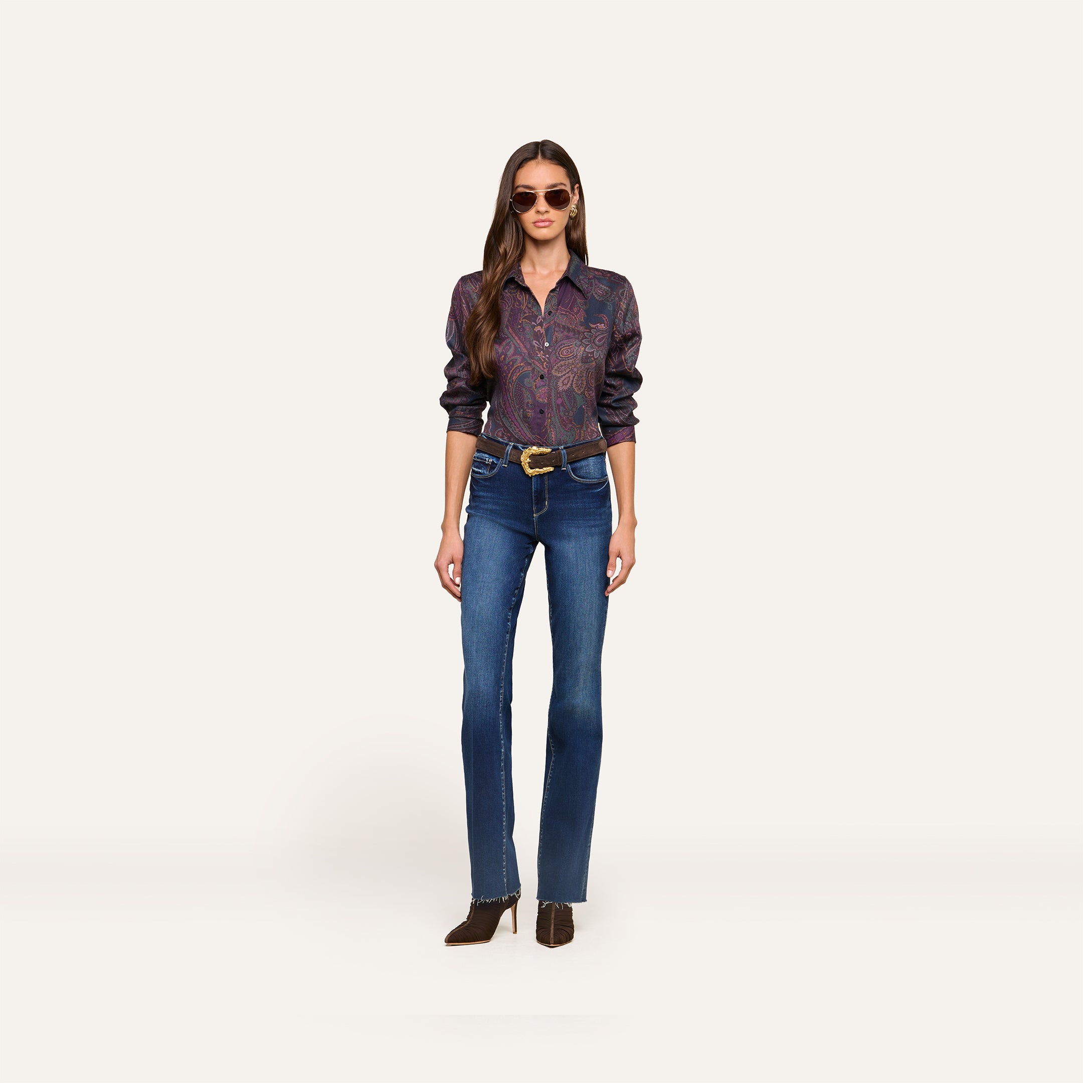 A woman wearing sunglasses, the Ellery Button-Down Shirt in a paisley print, blue jeans with a belt, and brown shoes stands against a plain white background.