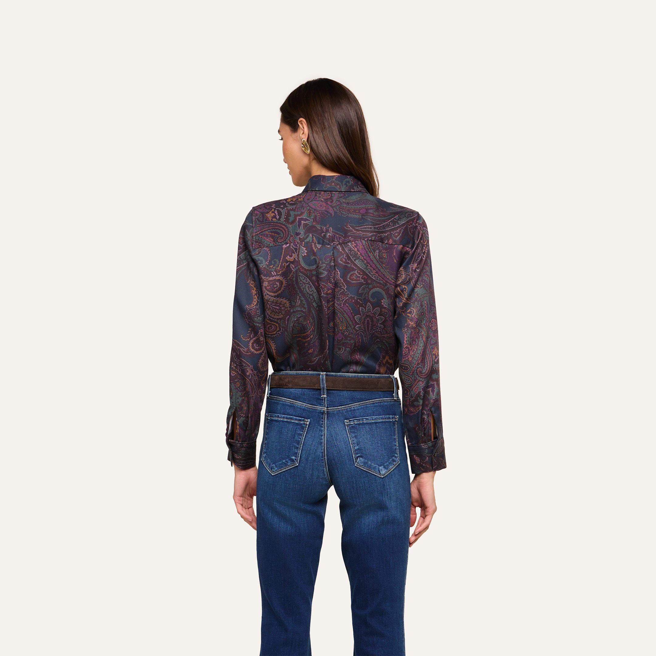 A woman stands with her back to the camera, wearing the Ellery Button-Down Shirt in a dark paisley print tucked into blue jeans, set against a plain light background.