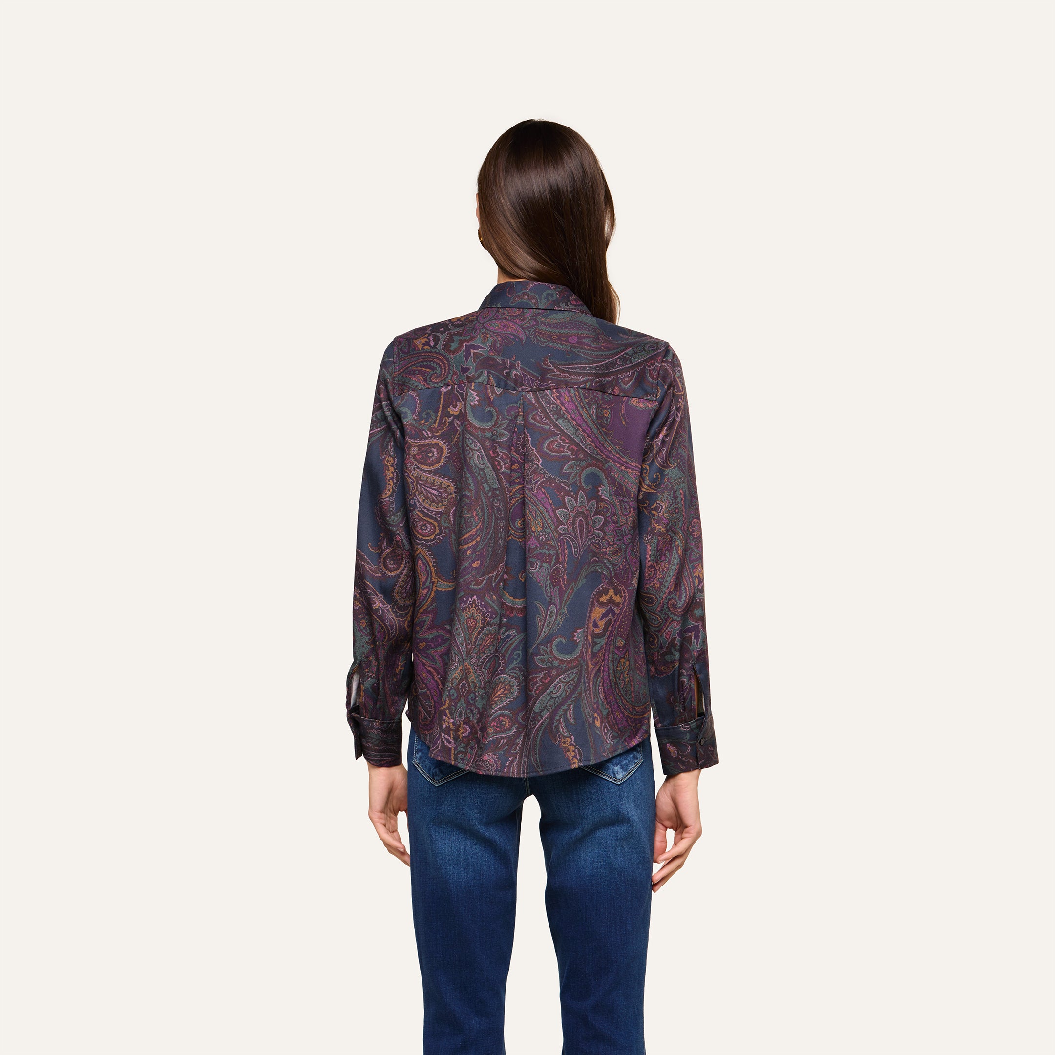 A person stands facing away, wearing the Ellery Button-Down Shirt—a dark paisley print blouse with long sleeves—paired with blue jeans against a plain, light background.