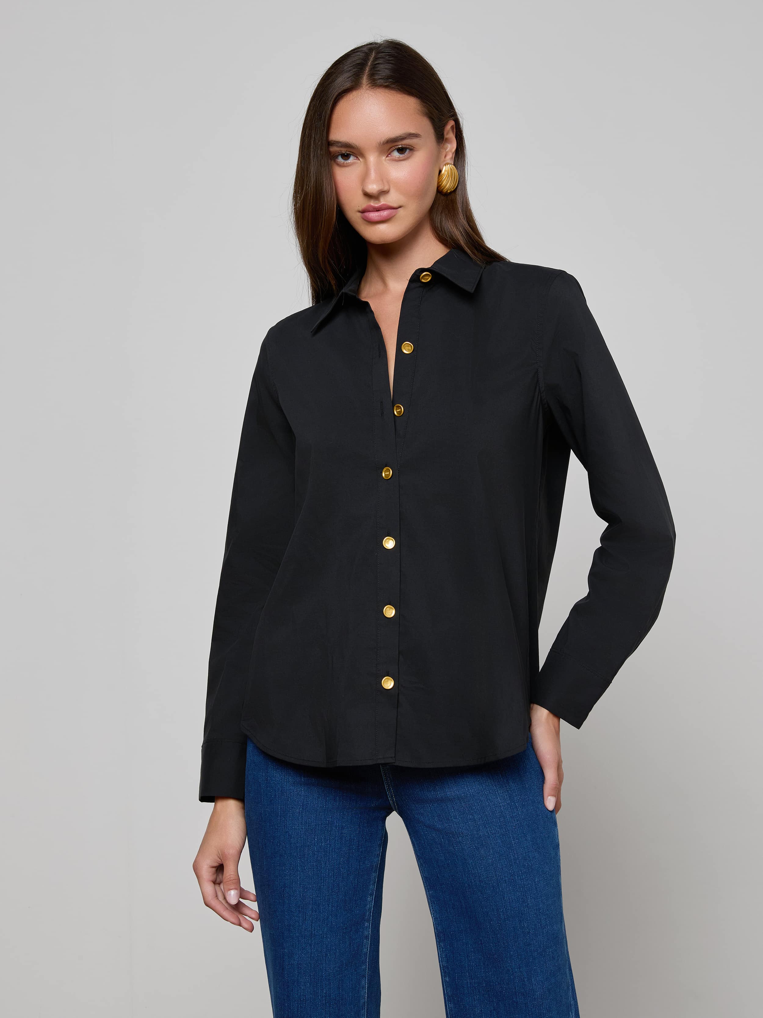 A woman wearing the Ellery Button-Down Shirt, a black stretch cotton blouse with gold buttons, paired with blue jeans stands against a plain light background, looking at the camera with one hand on her hip.