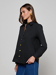Wearing the Ellery Button-Down Shirt, a woman with long brown hair styles it with blue jeans and large round earrings, standing against a plain light background.