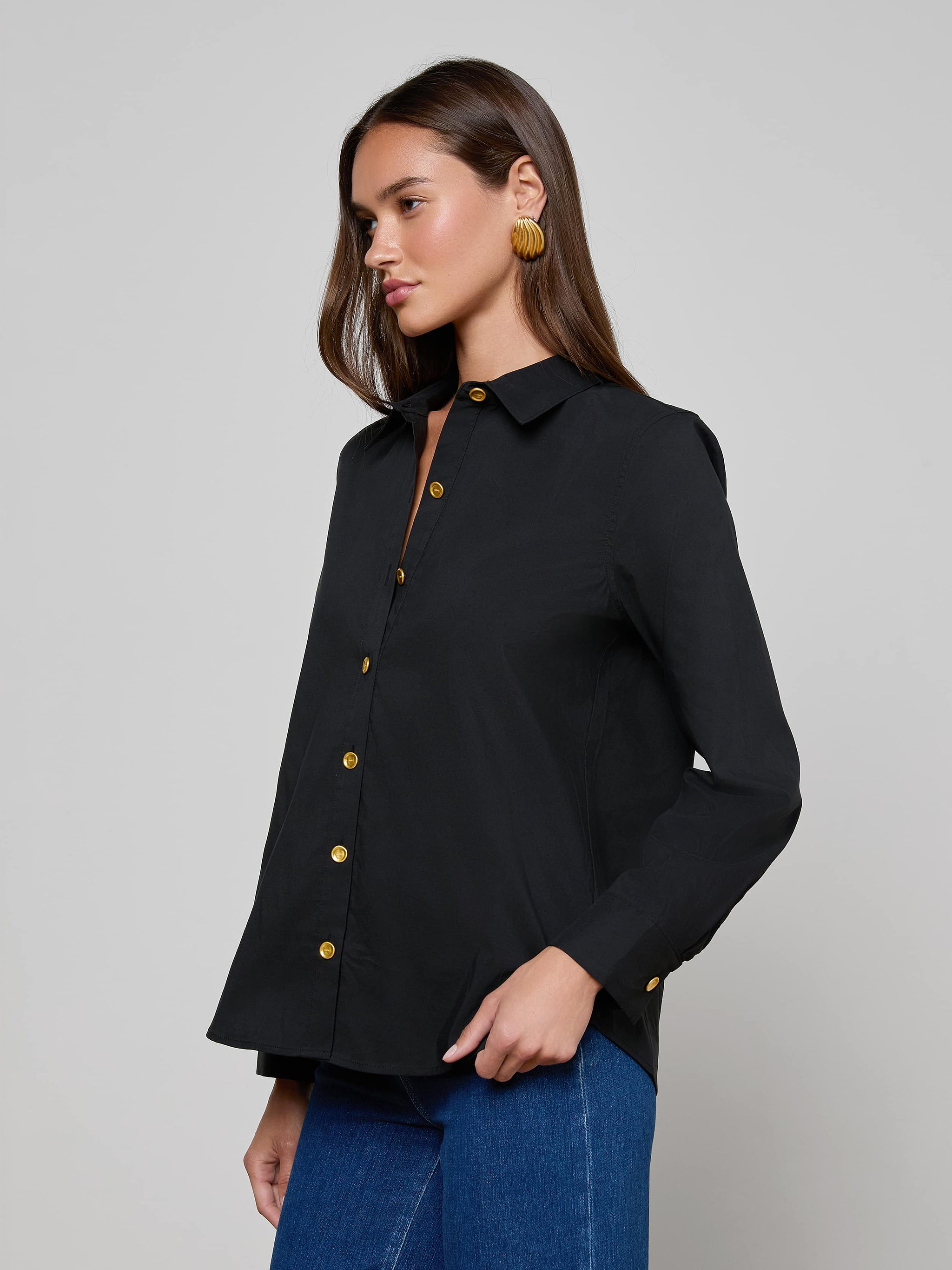 Wearing the Ellery Button-Down Shirt, a woman with long brown hair styles it with blue jeans and large round earrings, standing against a plain light background.
