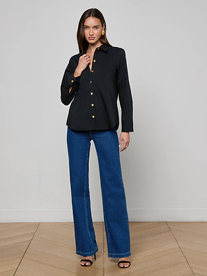 A woman with long brown hair poses indoors on a light wood floor against a gray wall, wearing the L'AGENCE Ellery Button-Down Shirt in black stretch cotton poplin with gold buttons, blue flared jeans, black heeled sandals, and gold earrings.