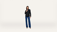 A woman stands against a plain white background, wearing a black blazer, dark blue wide-leg jeans, and black sandals.