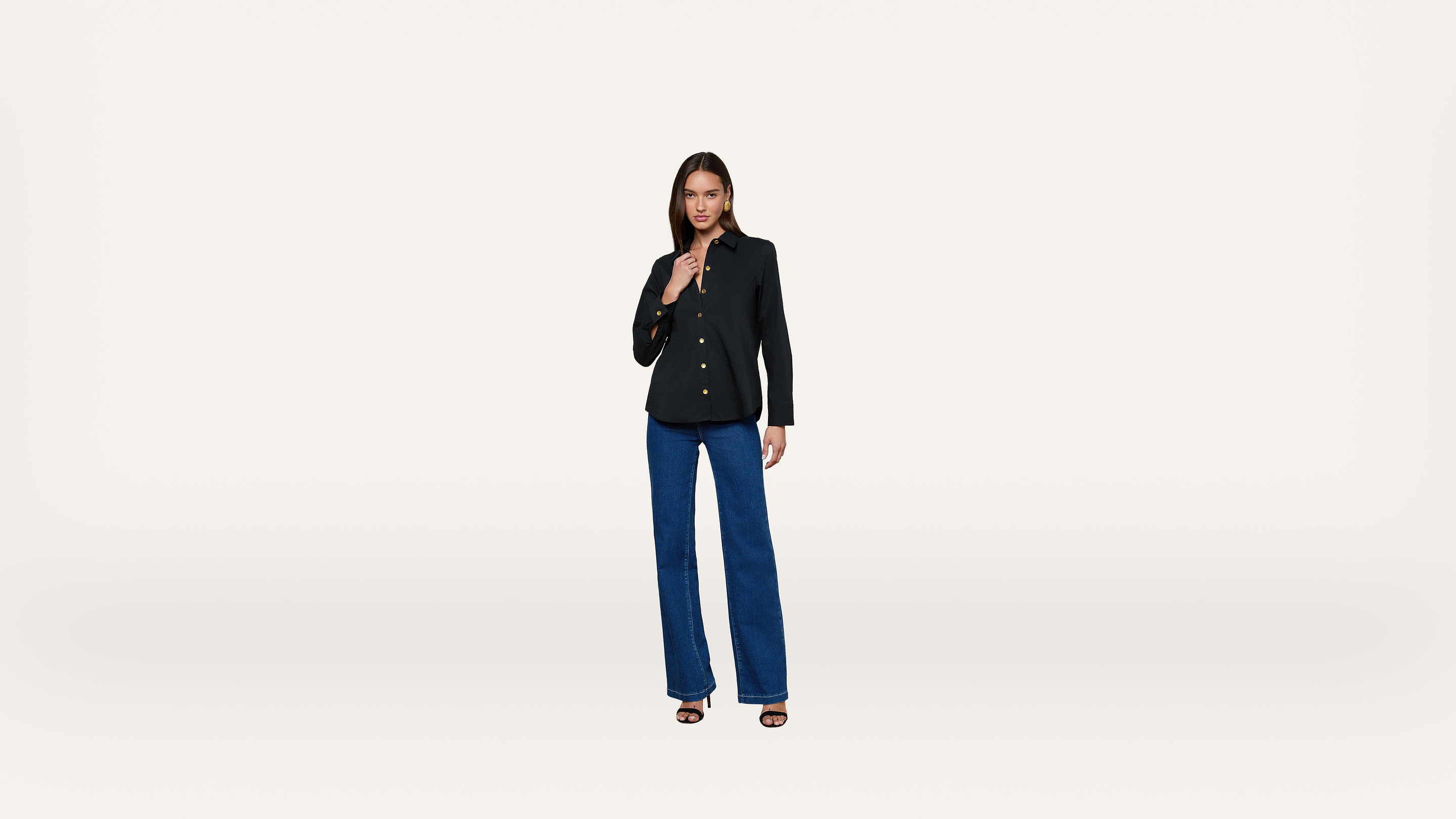 A woman stands against a plain white background, wearing a black blazer, dark blue wide-leg jeans, and black sandals.