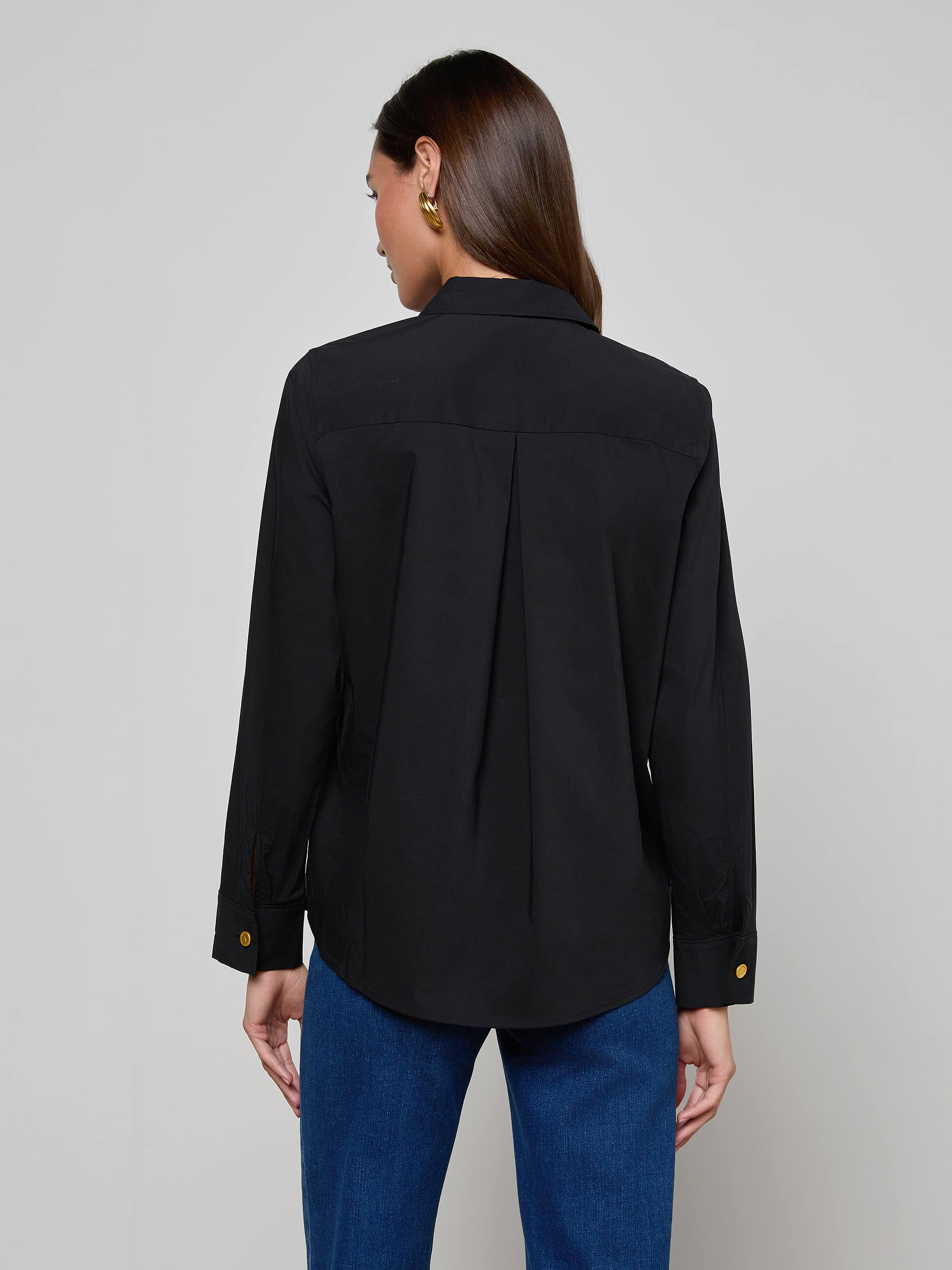 A woman with long brown hair is shown from the back against a plain light background, wearing the Ellery Button-Down Shirt in black stretch cotton and blue jeans.