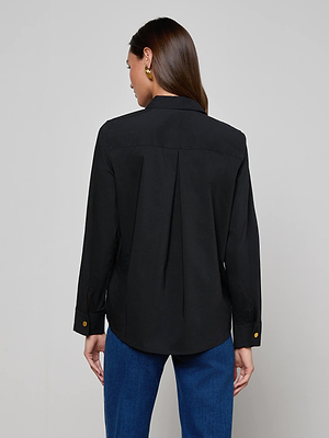 A woman with long brown hair models the L'AGENCE Ellery Button-Down Shirt in black stretch cotton poplin and blue jeans, standing with her back to the camera against a plain light gray background.