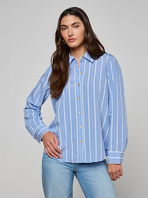 A woman with long brown hair stands against a light gray background, wearing the Ellery Poplin Shirt in relaxed fit blue and white stripes, paired with light blue jeans and one hand on her hip.