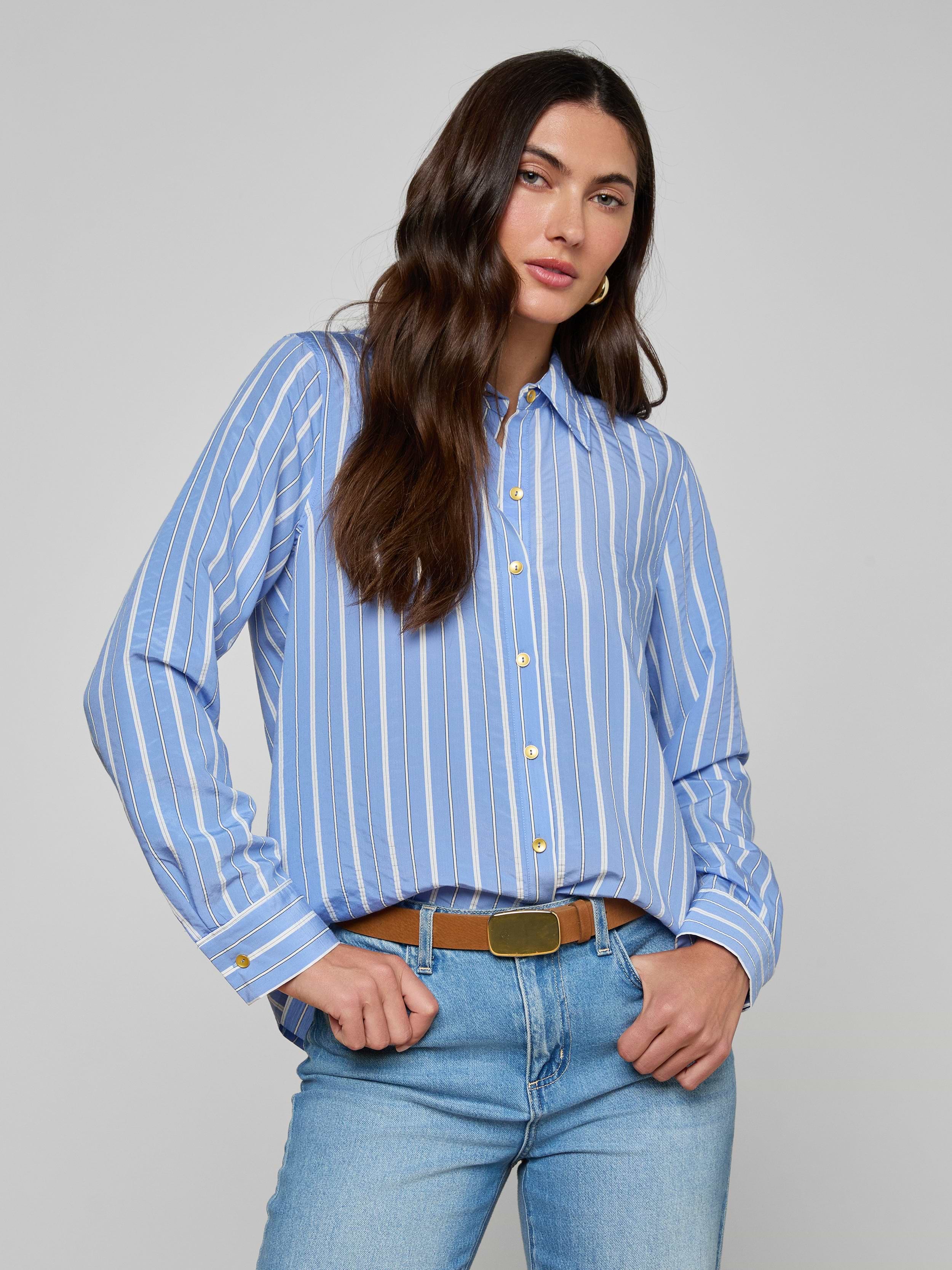 A woman with long dark hair stands against a plain background, hands in her pockets, wearing the Ellery Poplin Shirt— a blue and white striped button-down, tucked into light blue jeans.