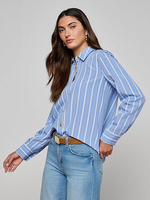 A woman with long brown hair wears the Ellery Poplin Shirt—blue and white striped, relaxed fit—paired with light blue jeans and a brown belt, standing against a plain light gray background.