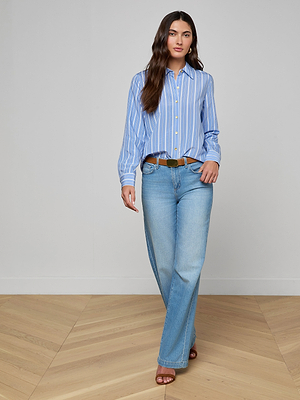 A woman with long brown hair and a neutral expression walks indoors on wooden flooring, wearing the Ellery Poplin Shirt blue and white stripes, light blue wide-leg jeans, a brown belt, and heeled sandals.