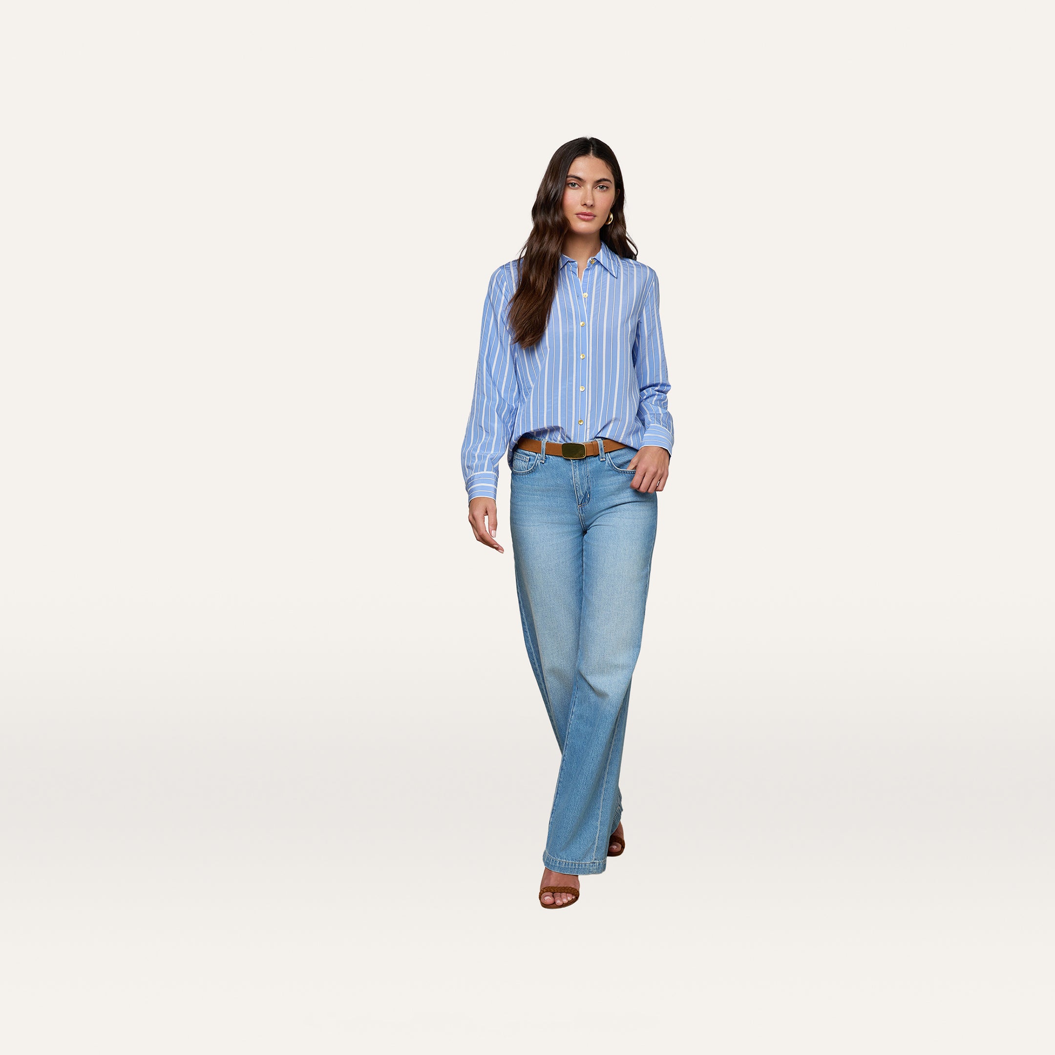 A woman stands facing forward wearing the Ellery Poplin Shirt—a relaxed fit blue striped button-down—paired with light blue wide-leg jeans, a brown belt, and brown sandals against a plain white background.
