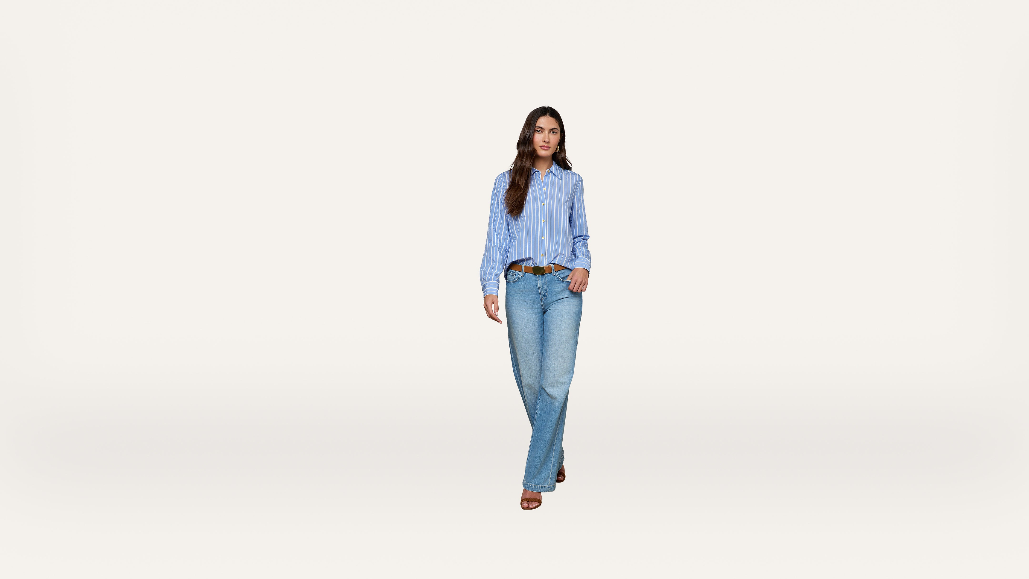 A woman wearing a blue striped shirt, light blue jeans, brown belt, and brown sandals walks toward the camera on a plain white background.